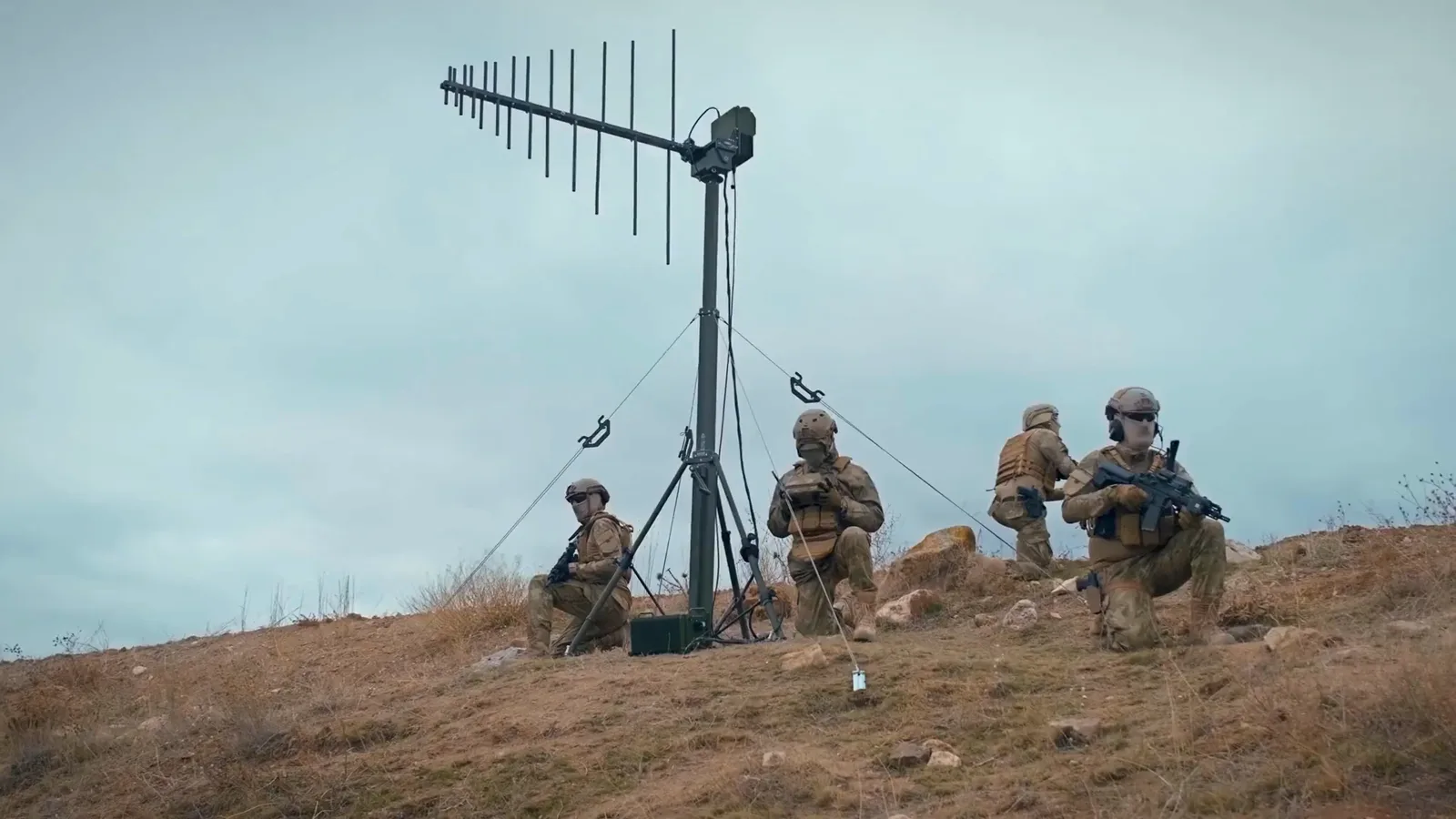 Guyed communications mast with a yagi array on an overcast hilltop, operators working at its base.